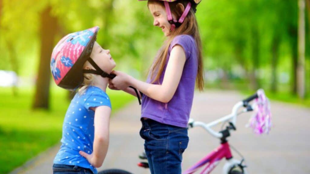 Two young girls wearing helmets and smiling, enjoying a sunny day at the park with a bicycle nearby.