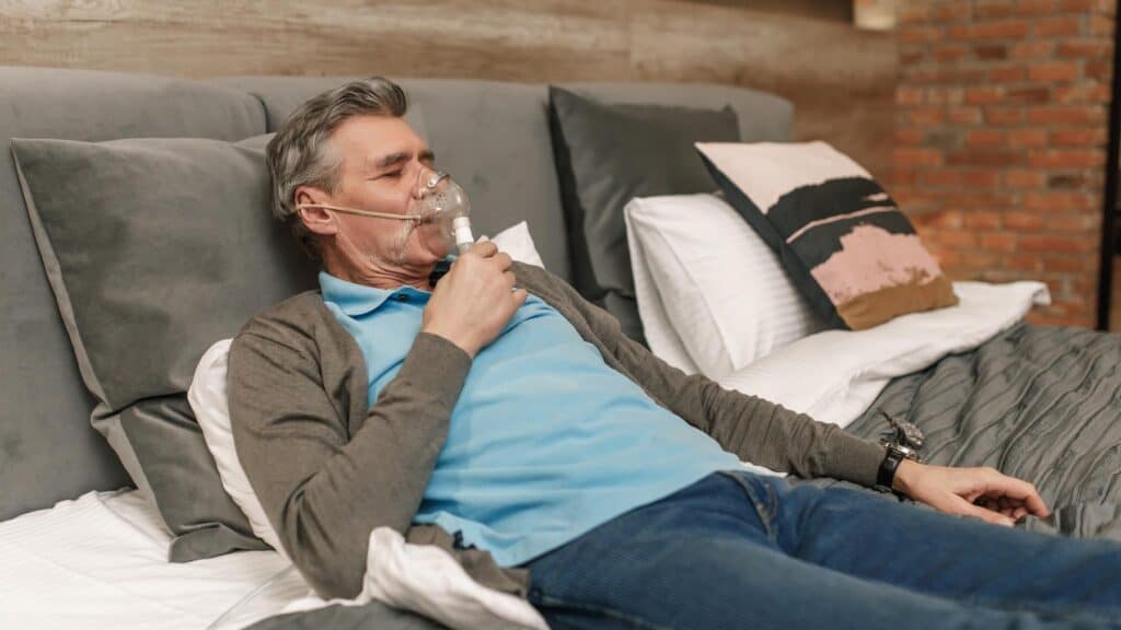 Elderly man using oxygen mask while resting on bed in a cozy bedroom setting.